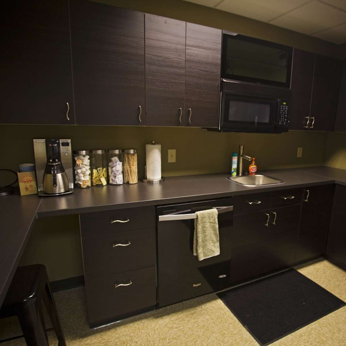 A modern kitchen with dark cabinetry, a countertop, a microwave, a coffee maker, and a refrigerator.