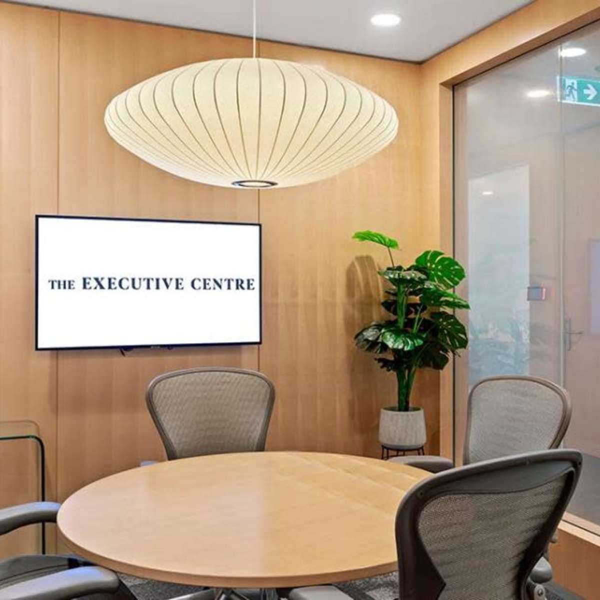 A minimalist meeting room features a round wooden table, ergonomic chairs, a wall-mounted television displaying "THE EXECUTIVE CENTRE," and a large pendant light above.