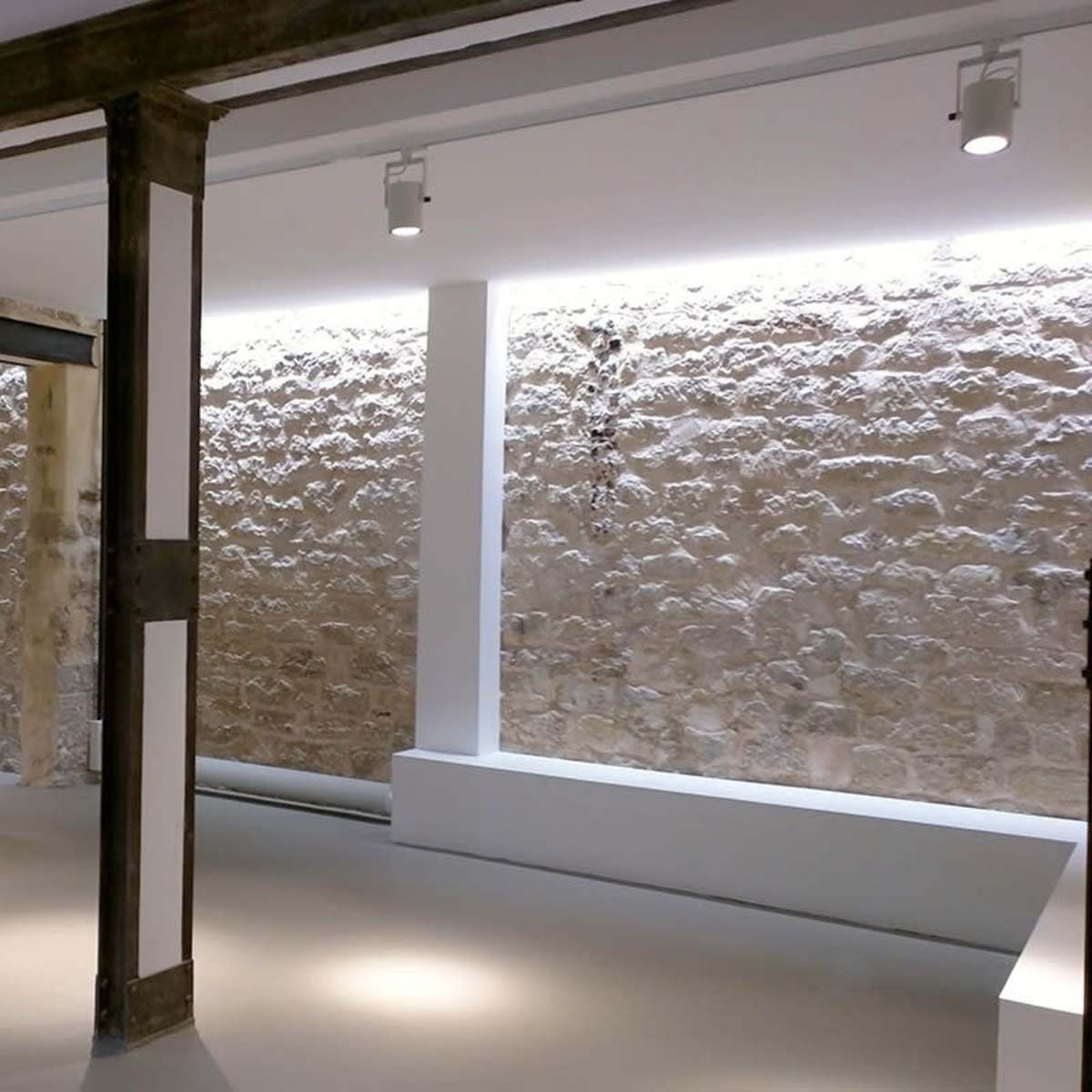 An interior space featuring exposed stone walls, minimalist lighting, and structural columns separating sections of the room.