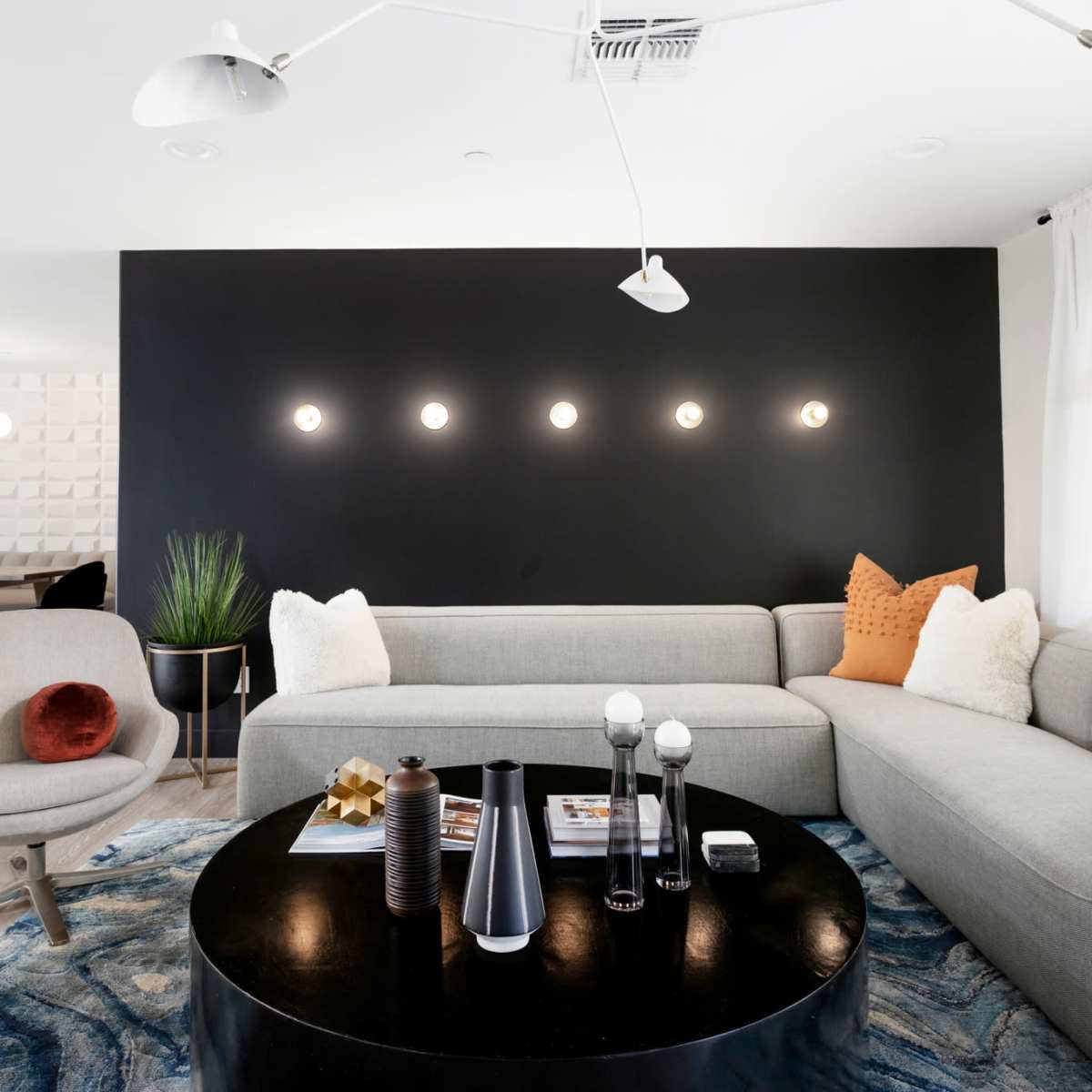 A modern living room features a gray sectional sofa, a round black coffee table, and a dark accent wall illuminated by five round lights.