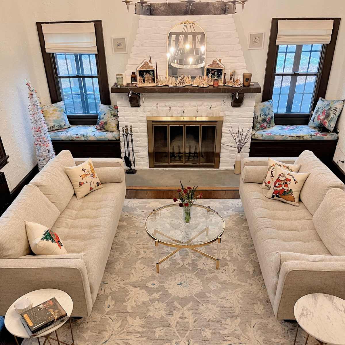 A cozy living room with two beige sofas facing each other, a round glass coffee table in the center, and a white-brick fireplace decorated with small houses and a Christmas tree.