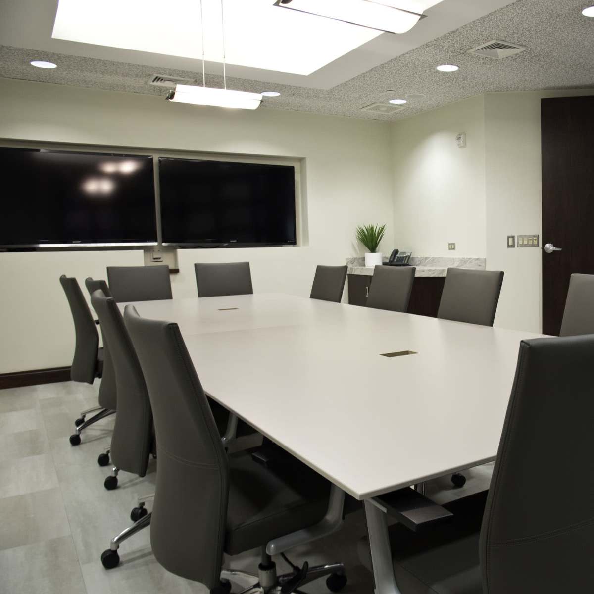 A modern conference room features a large white table surrounded by ergonomic chairs, with a wall-mounted screen and a glass-paneled door.