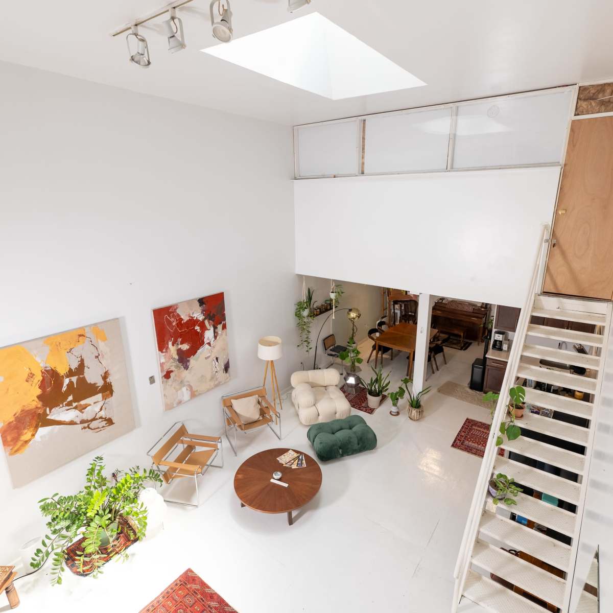 A bright, modern interior features a living area with art pieces, plants, and a staircase leading to an upper level.