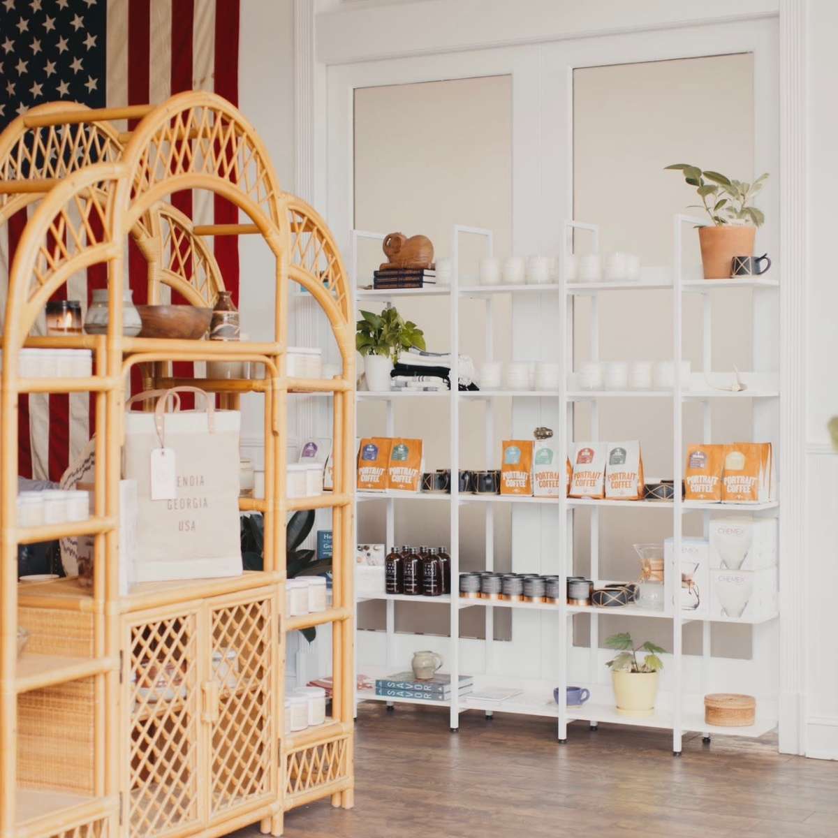 A bright interior features a wooden display shelf with woven accents, an American flag hanging on the wall, and a white shelving unit stocked with products.