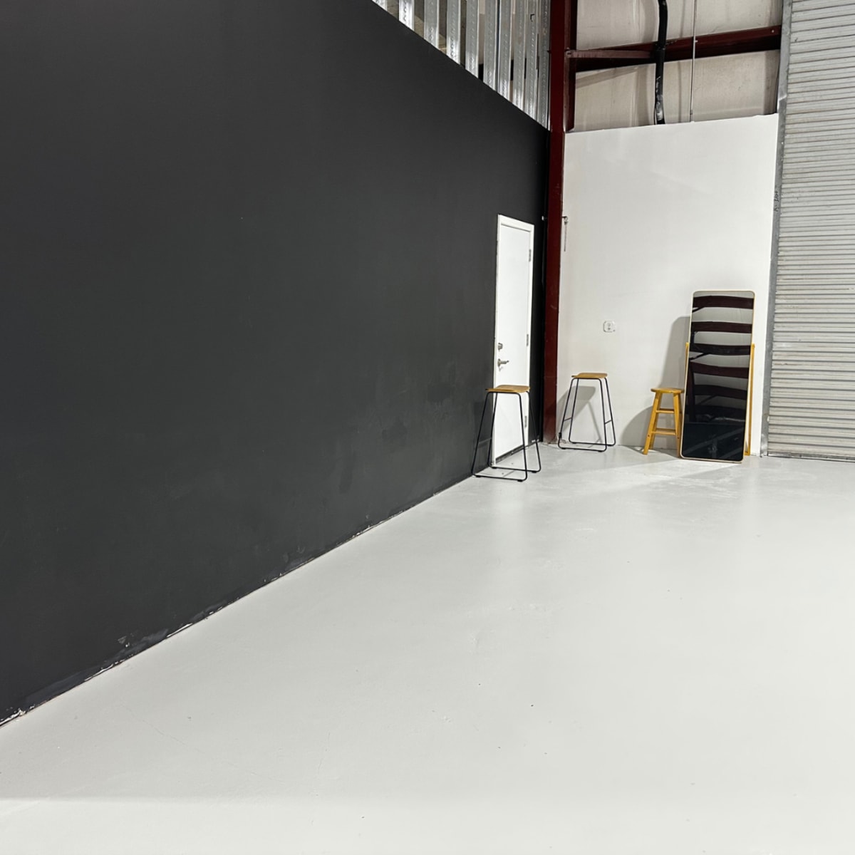 An interior space with a black wall, a white floor, a closed door, and two stools next to a mirror.