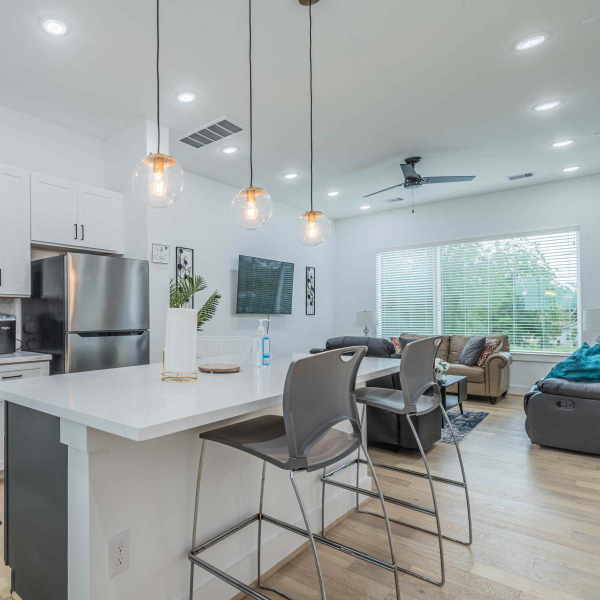 A modern kitchen with white cabinetry, stainless steel appliances, and a stylish countertop that opens into a living area with a large window and comfortable seating.