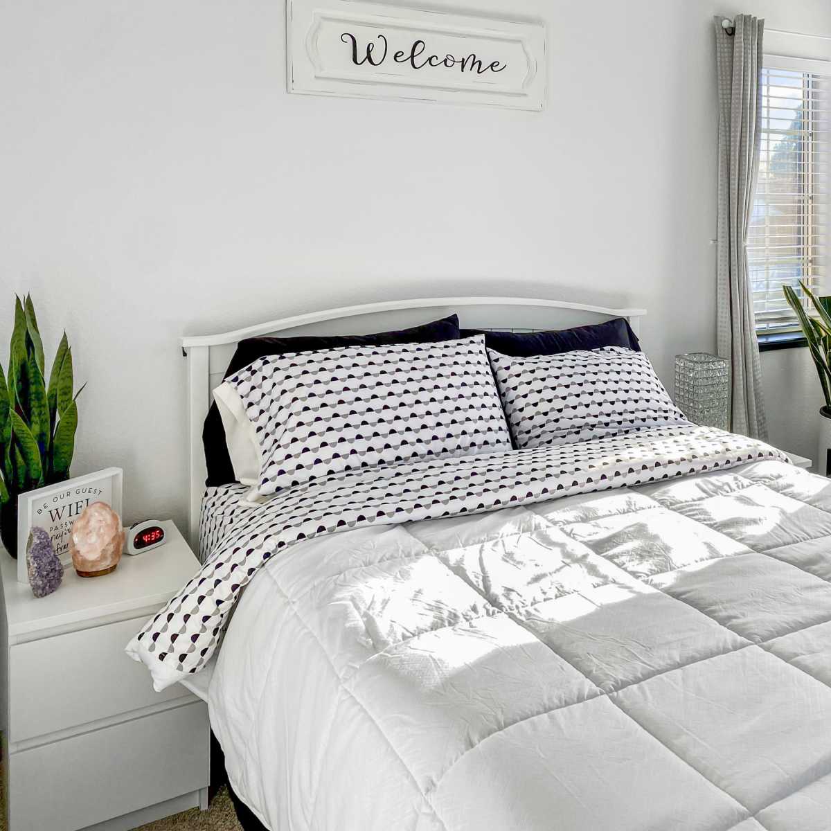 A neatly made bed with patterned pillows is positioned in a bright bedroom featuring a large window, potted plants, and decorative shelves.