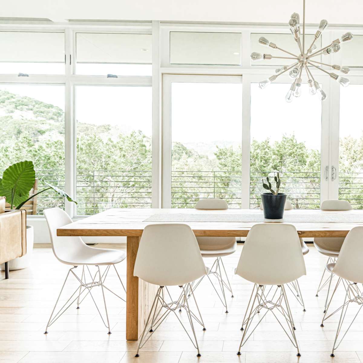 A long wooden dining table surrounded by white chairs is set against large windows overlooking greenery.