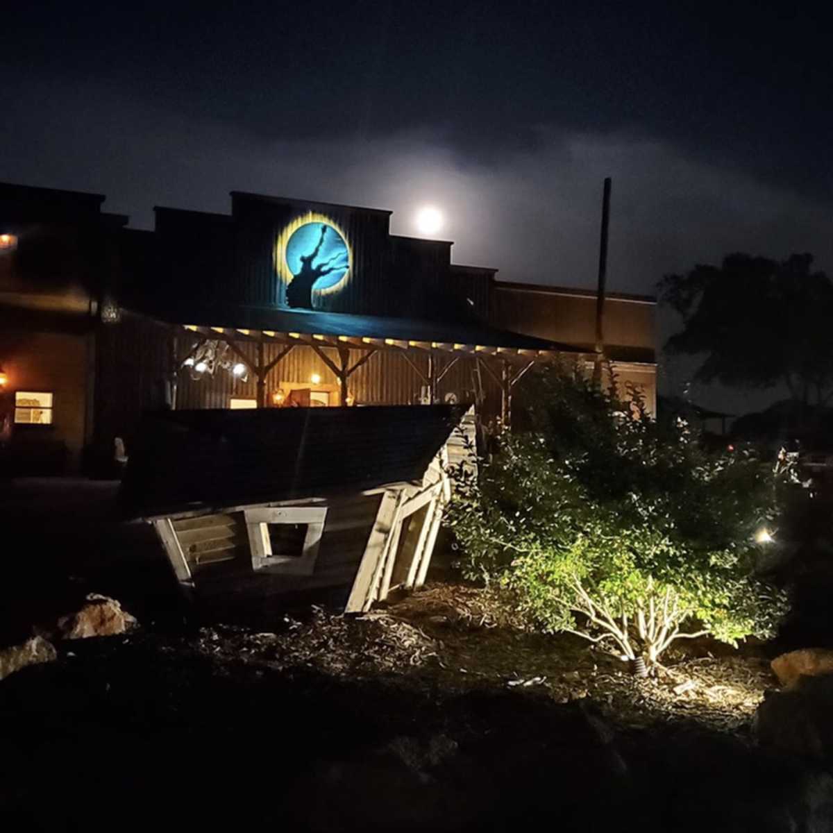 A full moon illuminates the facade of a rustic building, while a wooden sign and shrubbery are lit by ground lights in the foreground.