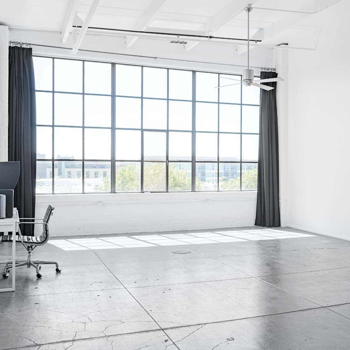 A bright, spacious room features a large window with a view, a ceiling fan, and minimal furniture including a chair and a stand.