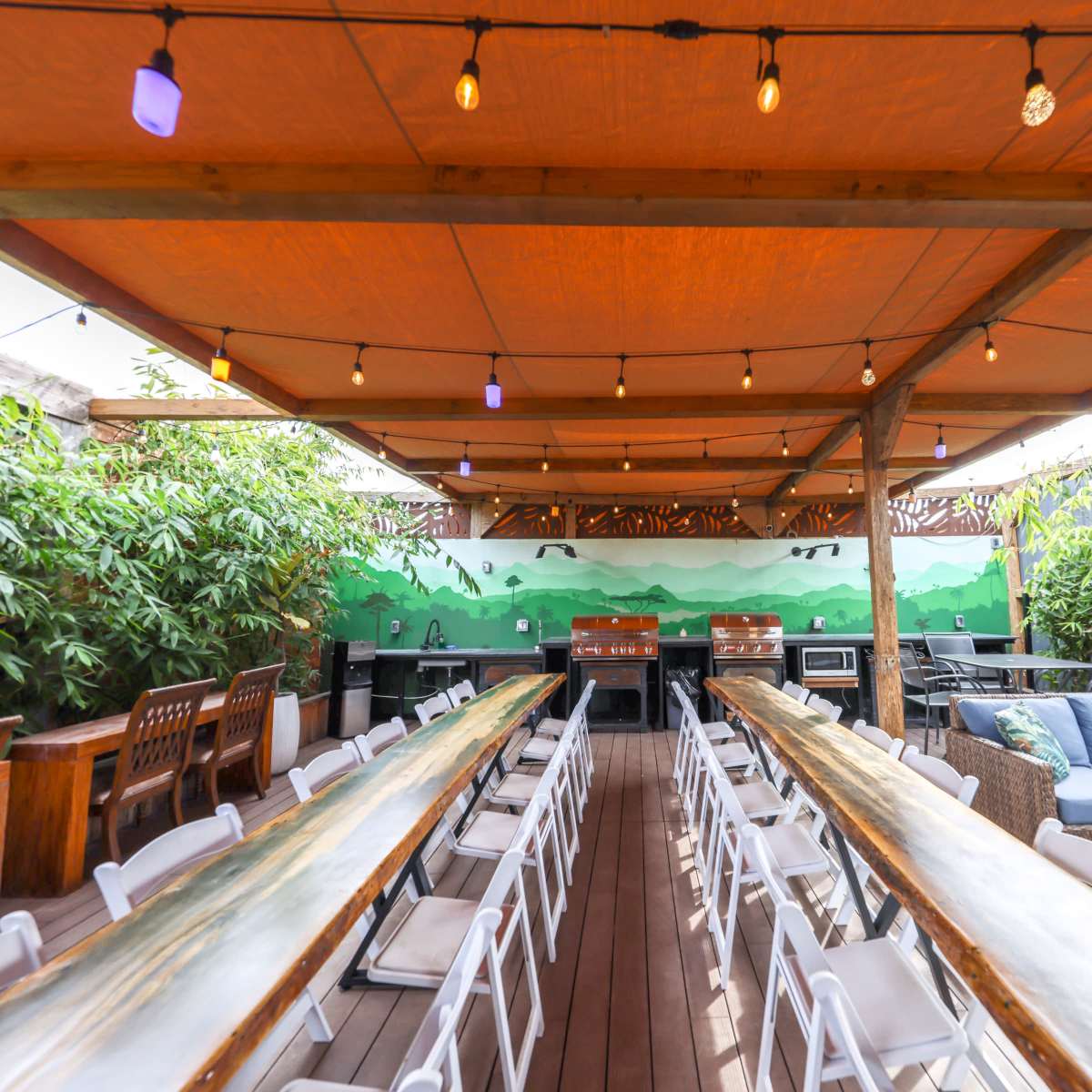 An outdoor dining space with long tables, string lights overhead, and a green mural on the wall surrounded by plants.
