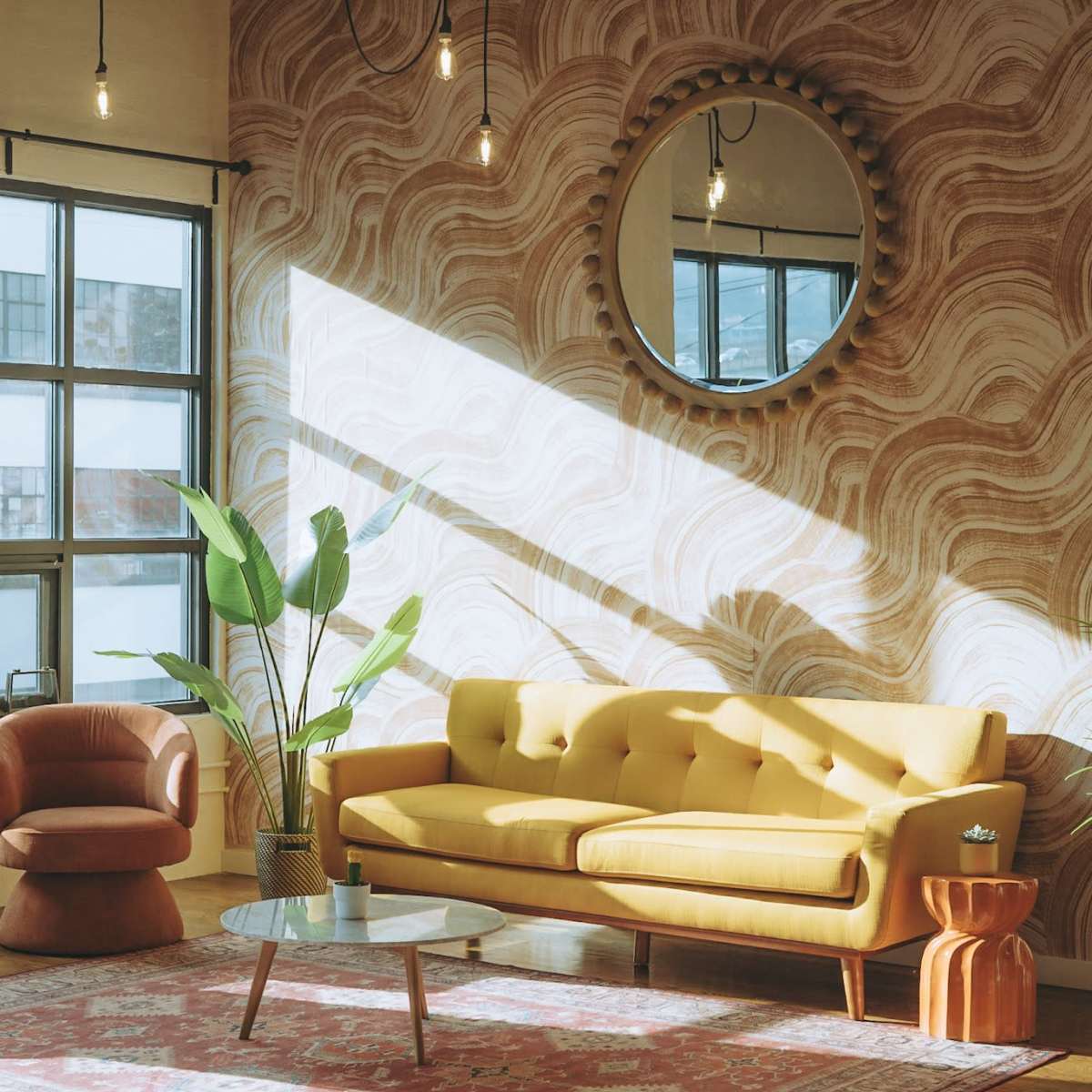 A cozy living room with a yellow couch, a round mirror, and a coffee table, illuminated by sunlight streaming through a large window.