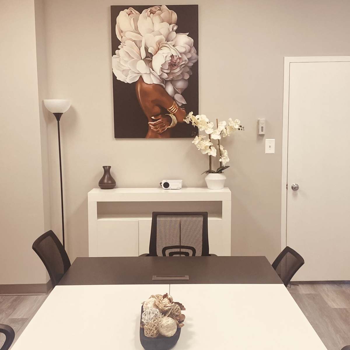 A modern meeting room features a central table surrounded by black chairs, with a large floral artwork on the wall, a white cabinet, and decorative elements including a vase of flowers.