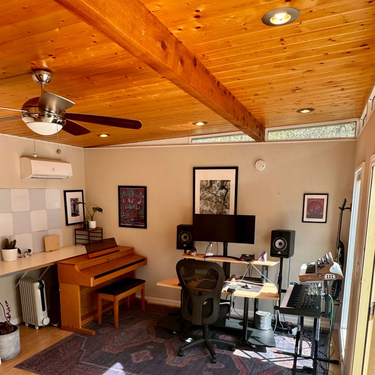 A bright room featuring a piano, a desk with a computer and audio equipment, and large windows allowing natural light to enter.