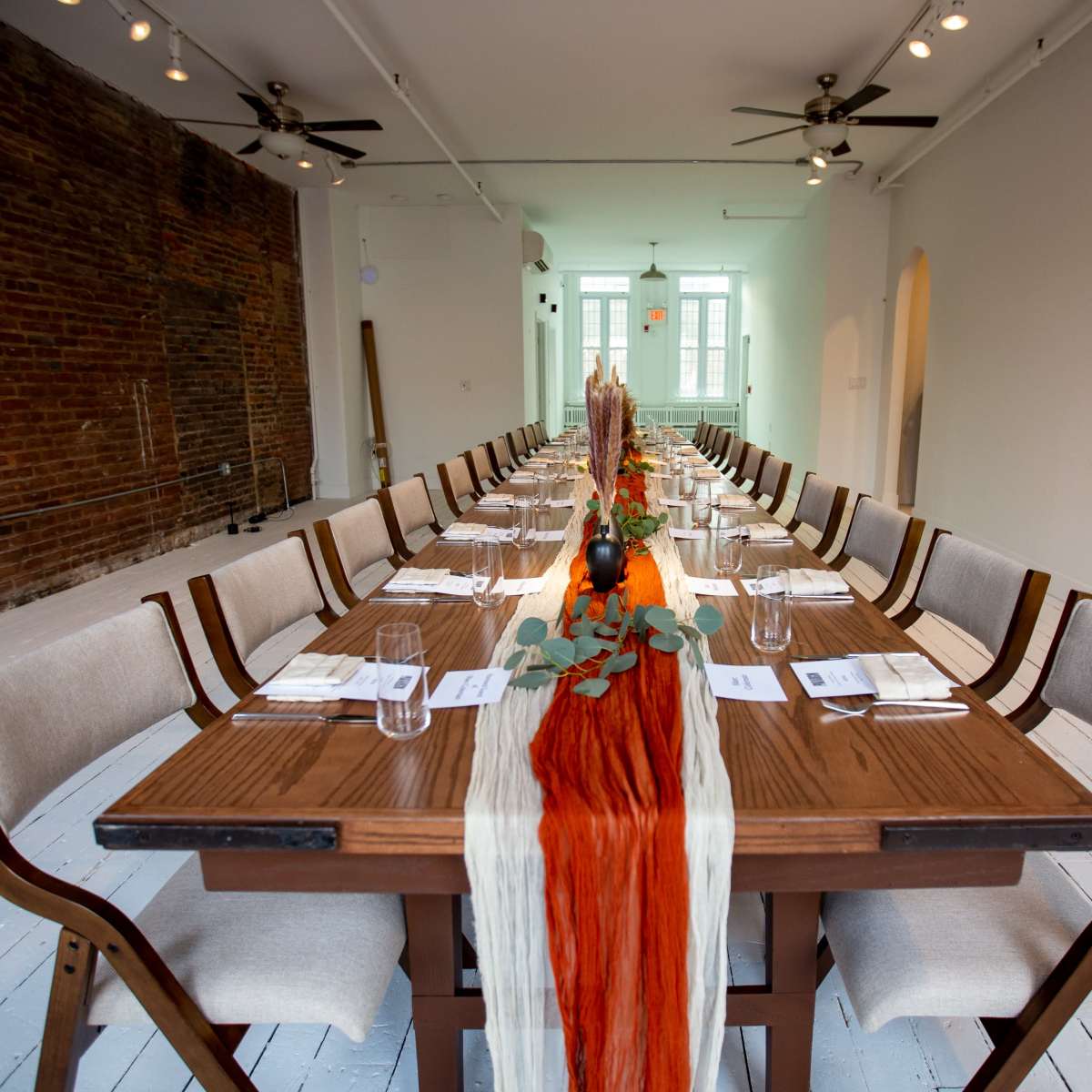A long wooden dining table is set with elegant tableware and a decorative runner, surrounded by chairs in a modern, well-lit room with exposed brick walls.