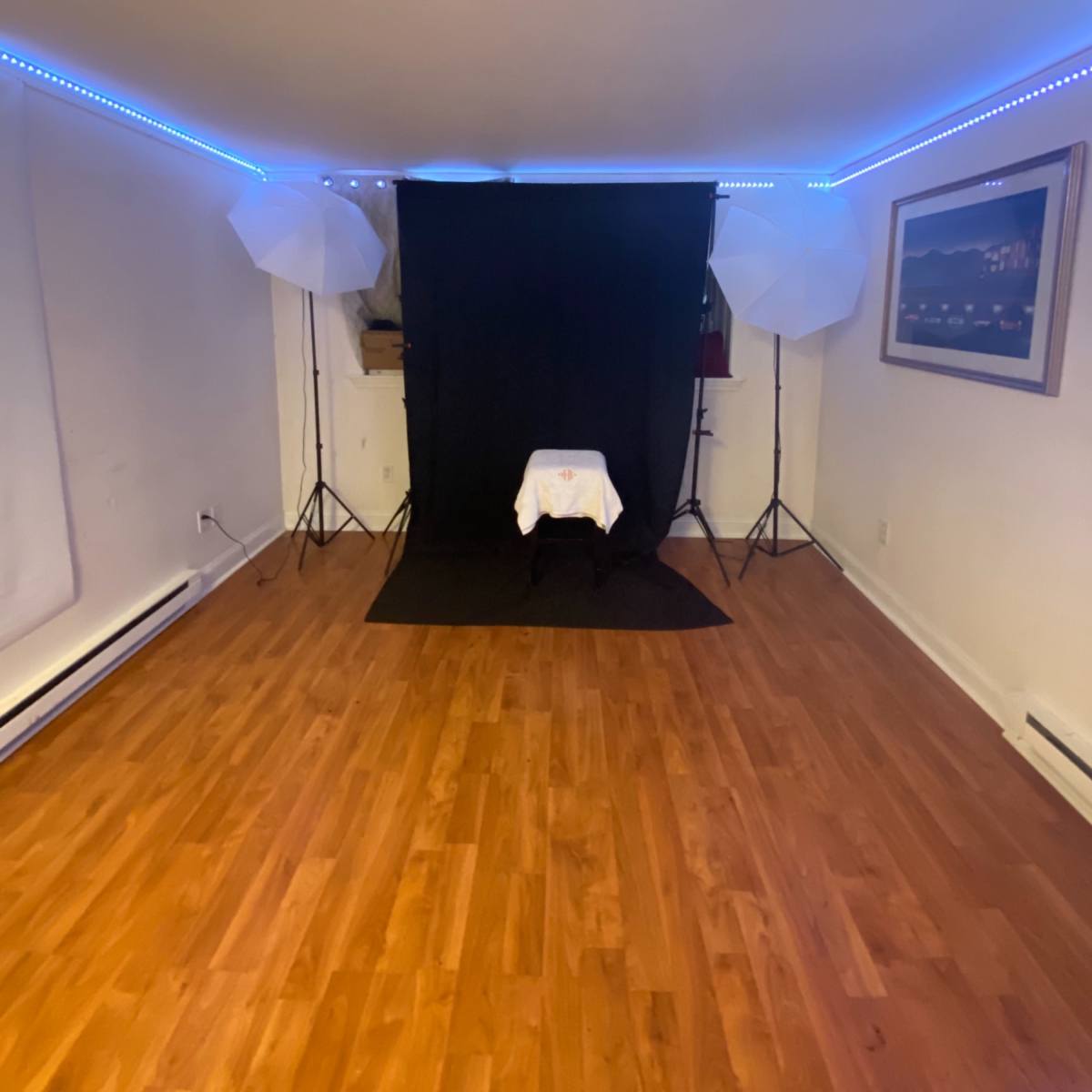 A photography studio setup with a black backdrop, two light stands with umbrellas, and a table covered with a white cloth in a room with wooden flooring.