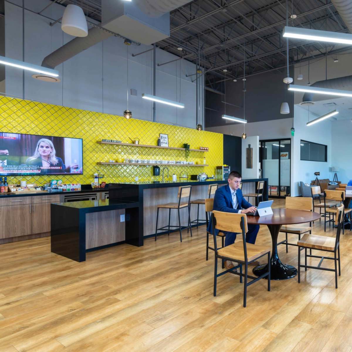 A modern workspace featuring a kitchen area with a yellow accent wall, several tables, and two individuals working on laptops.