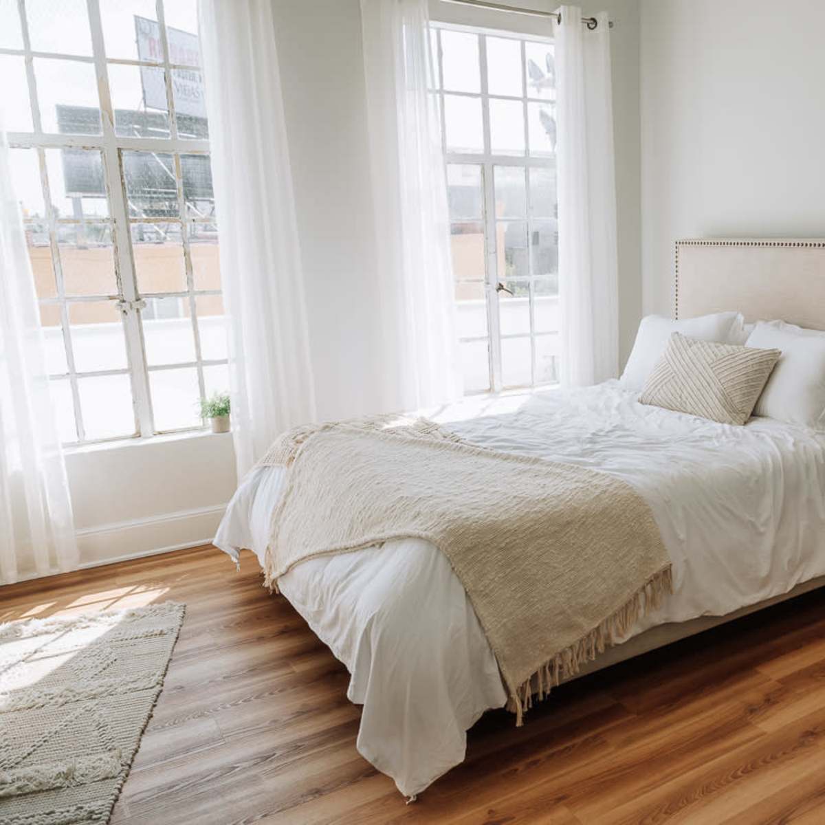 A bright bedroom features a bed with white bedding and a textured throw, complemented by large windows and wooden flooring.