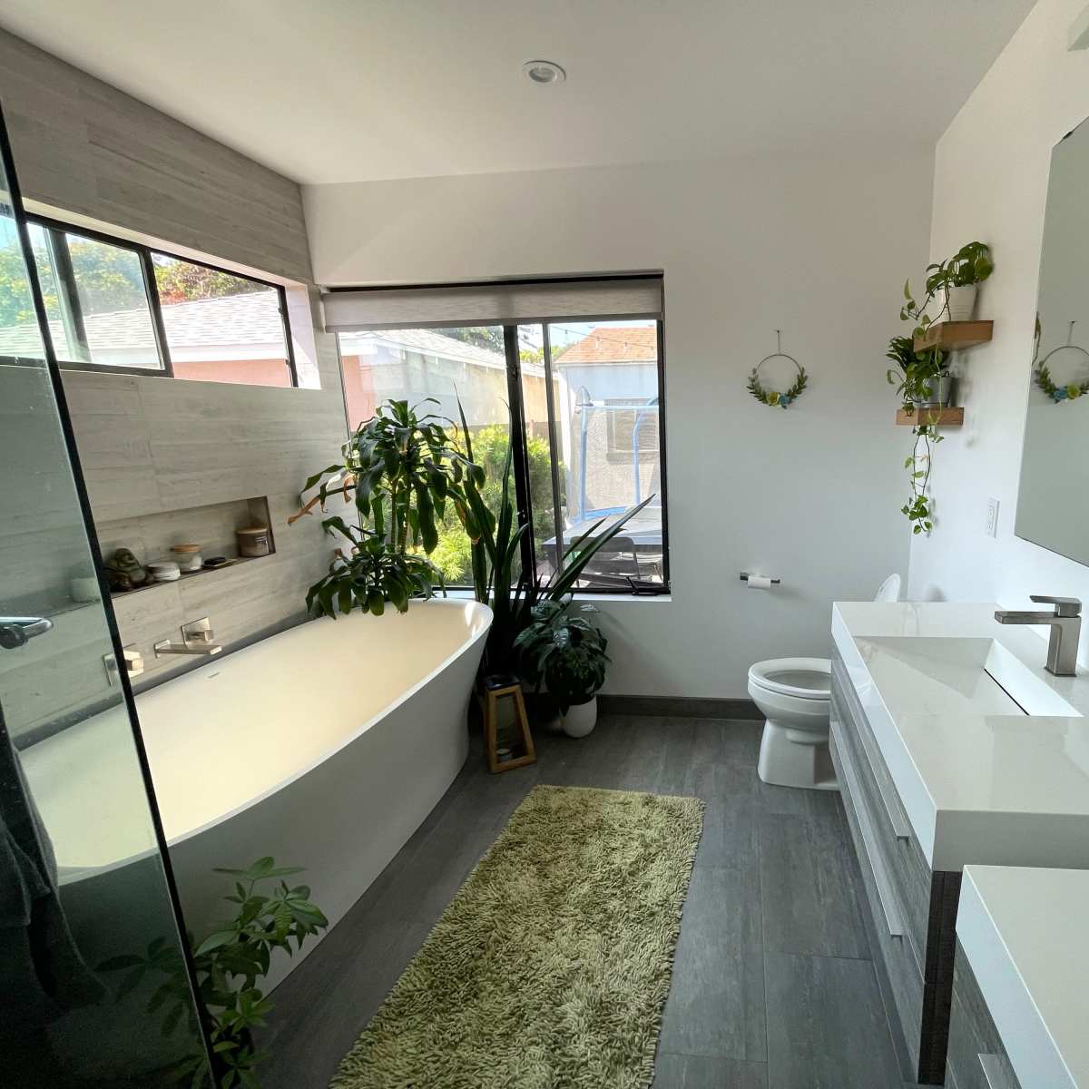 A modern bathroom featuring a freestanding bathtub, a large mirror above a vanity, and plants arranged around a window with natural light.