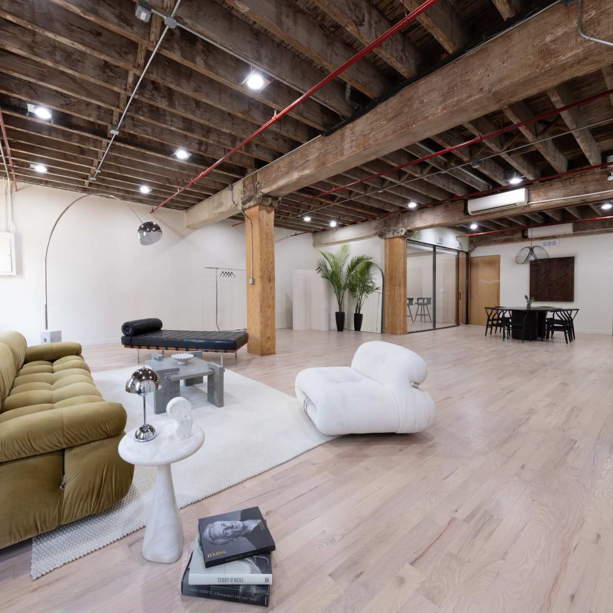 A spacious, open room with wooden beams, light-colored flooring, and modern furniture, including a green sofa, a coffee table, and a dining area in the background.