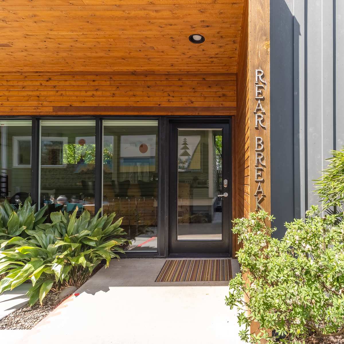 The entrance of a building with a wooden overhang, glass doors, and framed text that reads "REAR BREAK," surrounded by greenery.