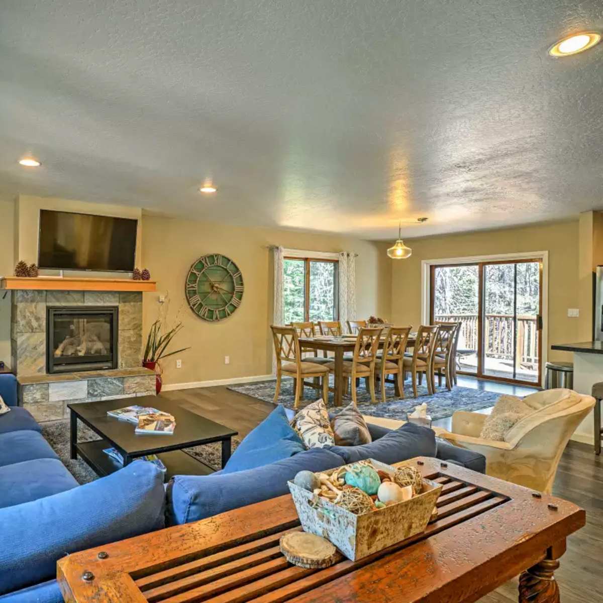 The living area features a blue sectional sofa, a coffee table, a stone fireplace, and a dining table with chairs next to a large window overlooking a deck.