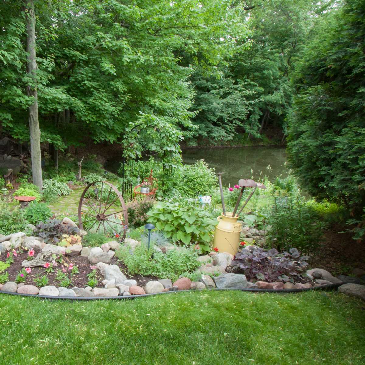 A landscaped garden features a winding path lined with stones, various plants, an old wheel, and a few colorful decorative items beside a pond.