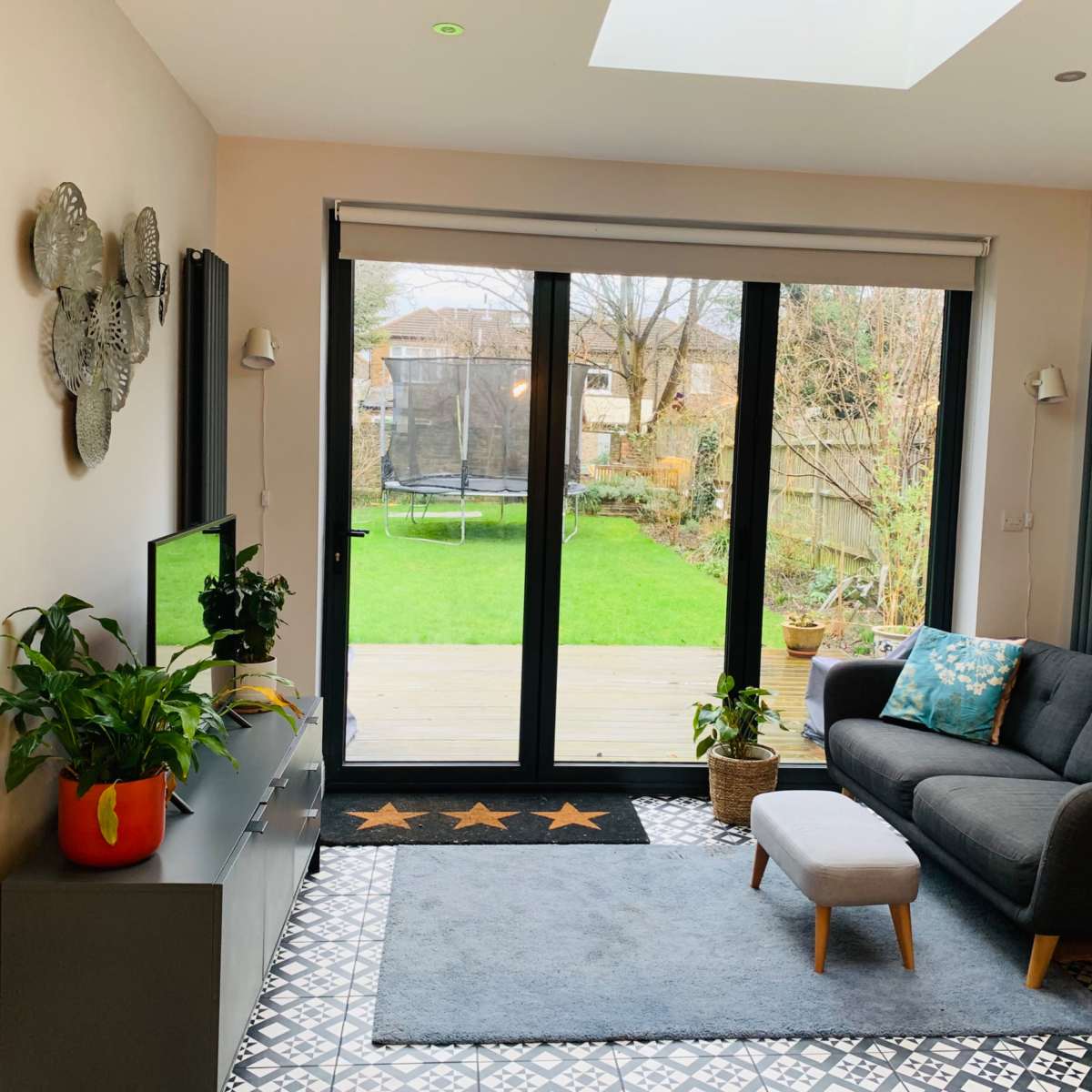 A modern living room with a gray sofa, a low coffee table, a television unit, and large glass doors leading to a garden.
