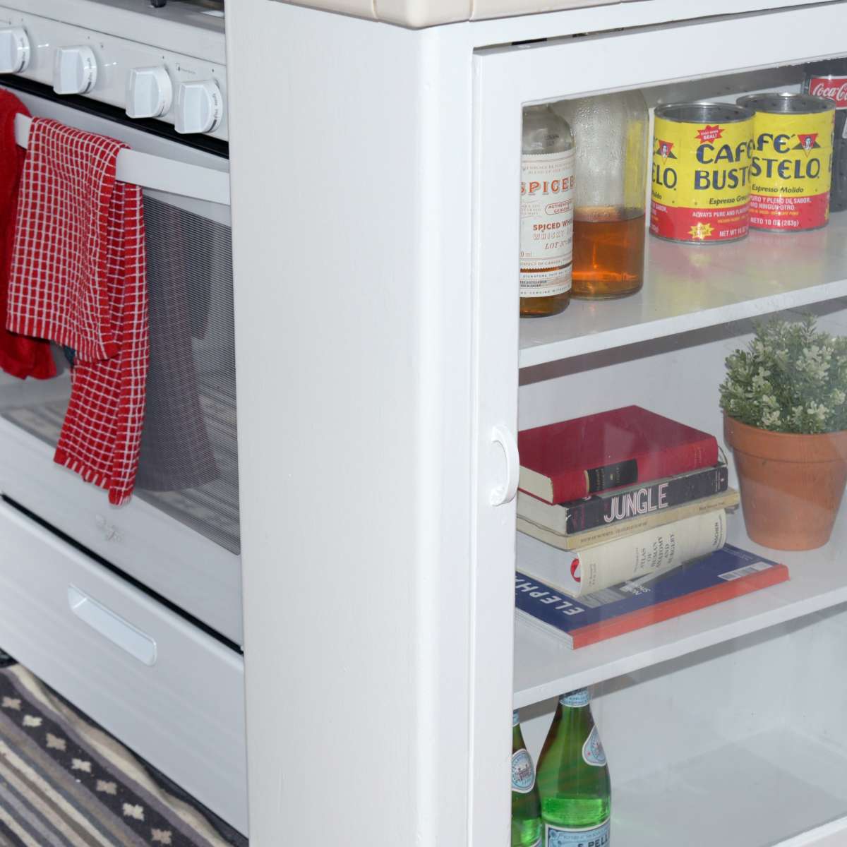 A white kitchen with an oven, dish towels hanging, and a glass-fronted cabinet displaying books, a potted plant, and drinks.
