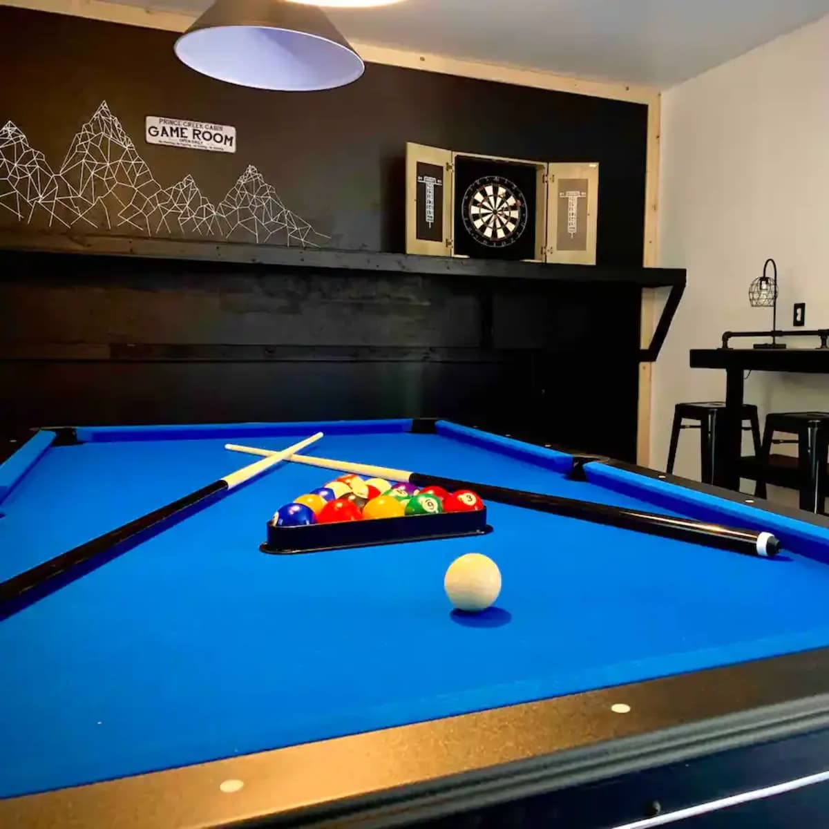 A game room featuring a blue pool table with balls arranged in a rack, a dartboard on the wall, and a bar-height table with stools.