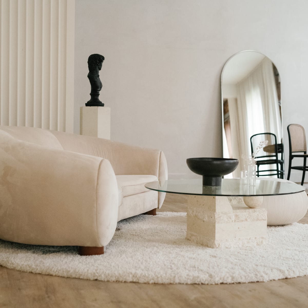 A plush beige sofa sits on a textured rug beside a round glass coffee table, with a sculptural bust and a mirror in the background.