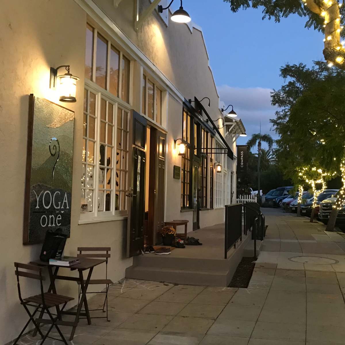 A yoga studio with string lights on trees is located along a sidewalk lined with parked cars in the evening.
