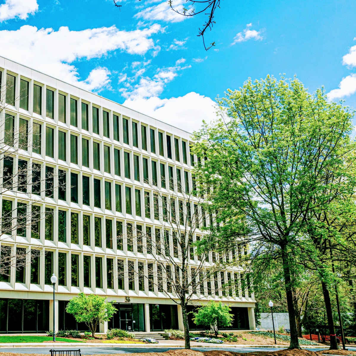 A modern office building with large windows stands among trees under a partly cloudy sky.