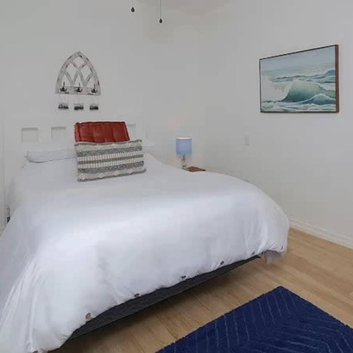 A bright bedroom featuring a large bed with white bedding, two bedside tables with lamps, and a painting of ocean waves on the wall.
