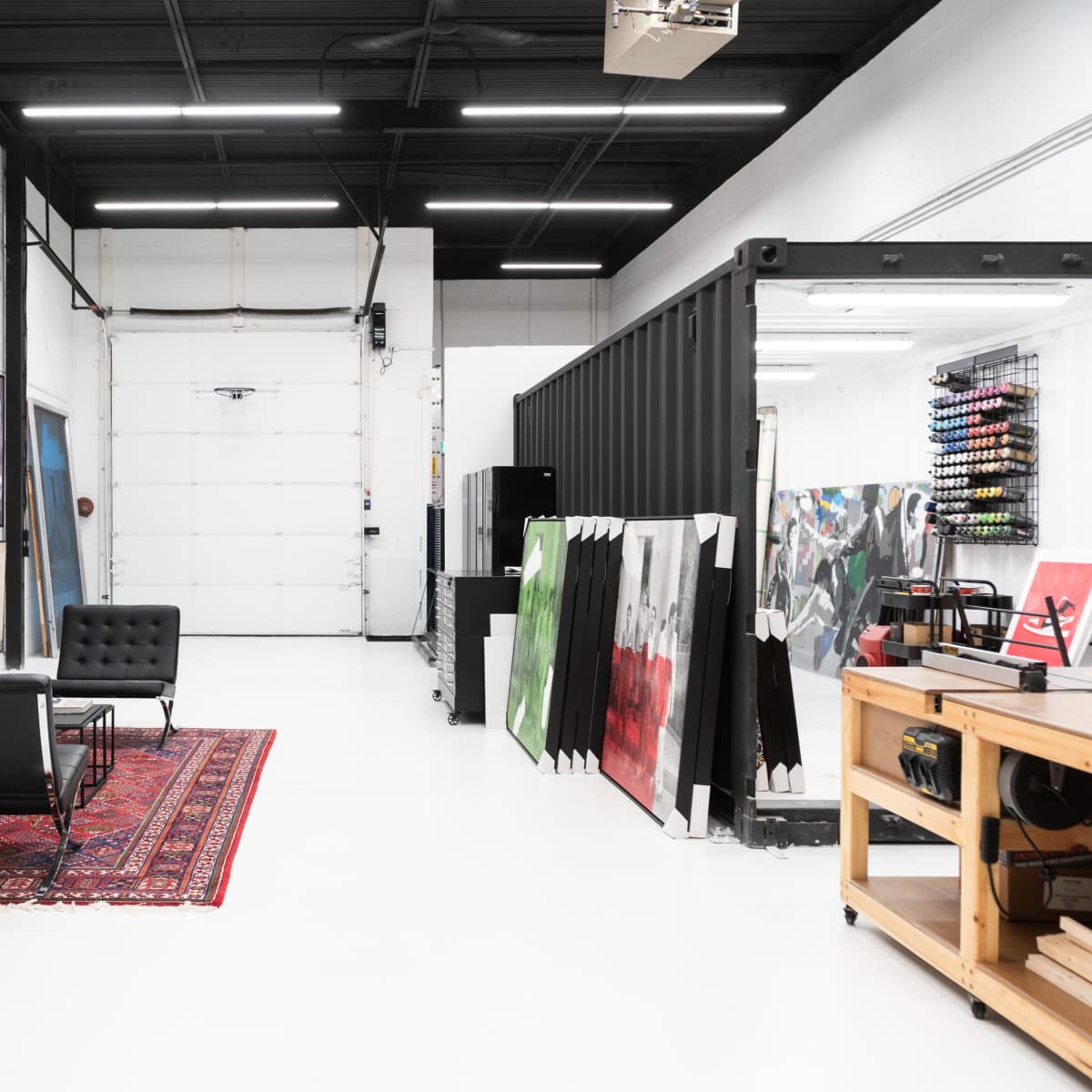 An industrial art studio with a seating area featuring a gray sofa and black chairs, surrounded by colorful artworks and a large garage door.