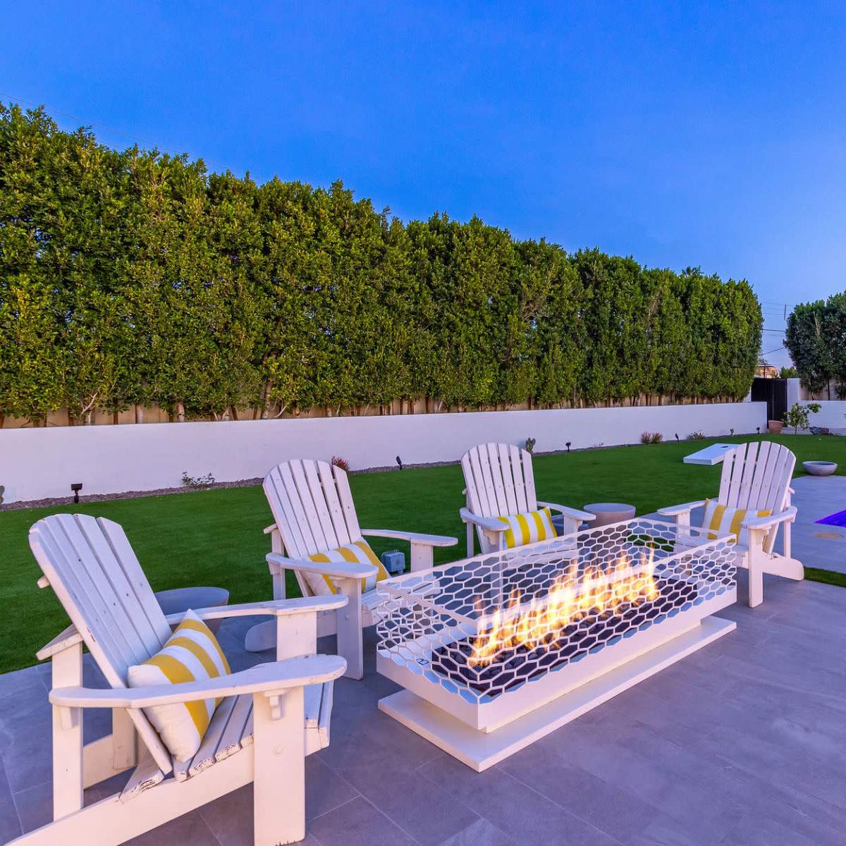 A backyard scene with five white Adirondack chairs arranged around a fire pit, a small swimming pool in the corner, and neatly trimmed hedges along a white wall.