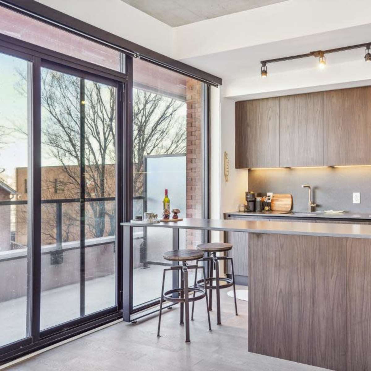 A modern kitchen features a wooden island, bar stools, and large glass doors opening to a balcony with trees visible outside.