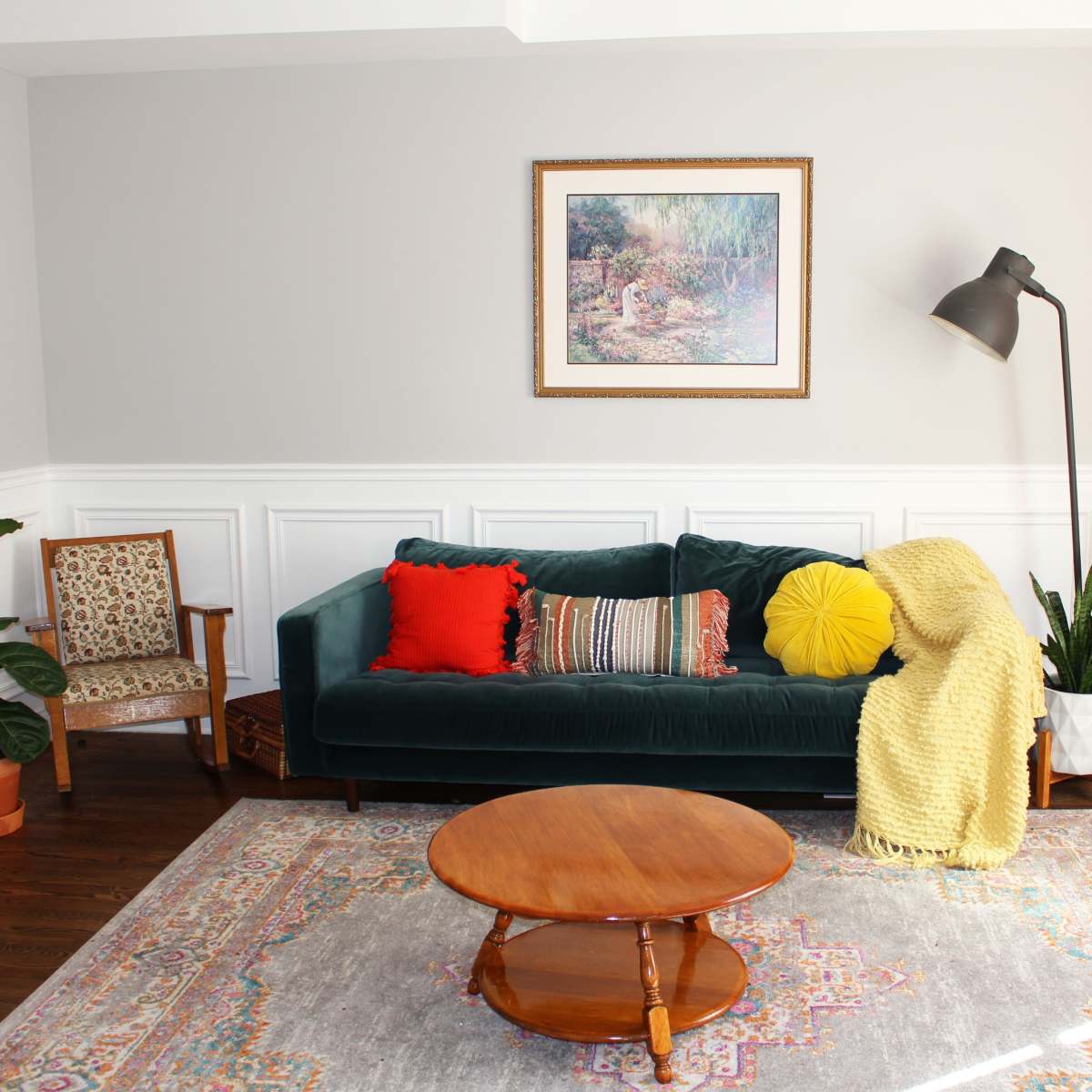 A living room features a green velvet sofa adorned with colorful pillows, a round wooden coffee table, a patterned area rug, and several plants alongside a floor lamp and framed artwork on the wall.