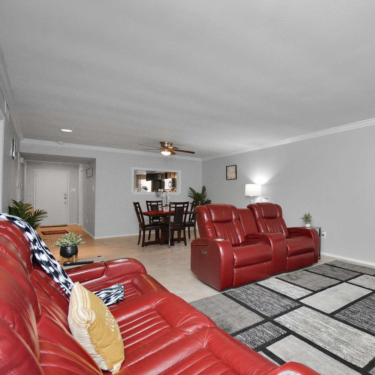 A spacious living room with red leather recliners, a flat-screen TV mounted on the wall, a patterned area rug, and a dining area visible in the background.