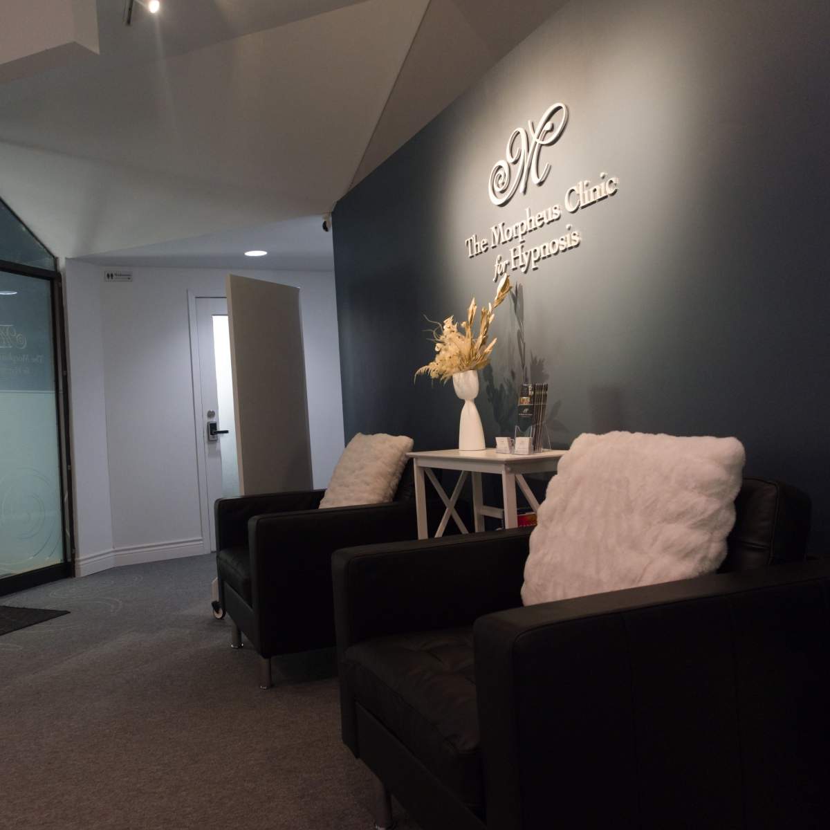 A modern reception area with two black chairs, a small table, and a sign on the wall displaying the clinic's name.