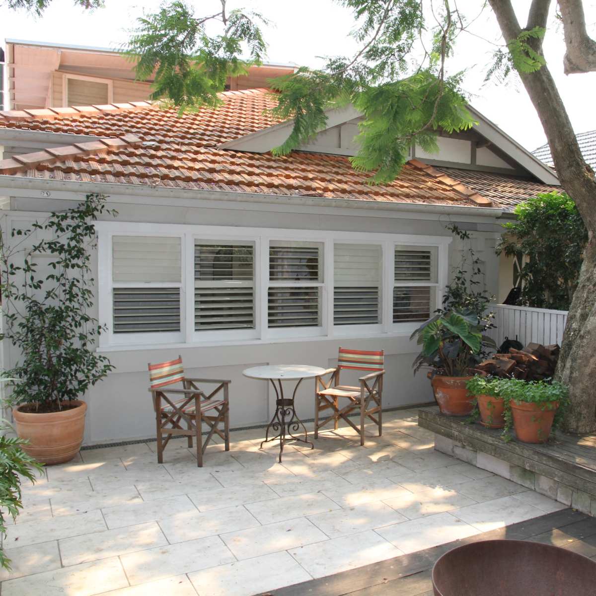 A patio area features two chairs and a small table beside a house with a tiled roof, surrounded by potted plants and a large tree.