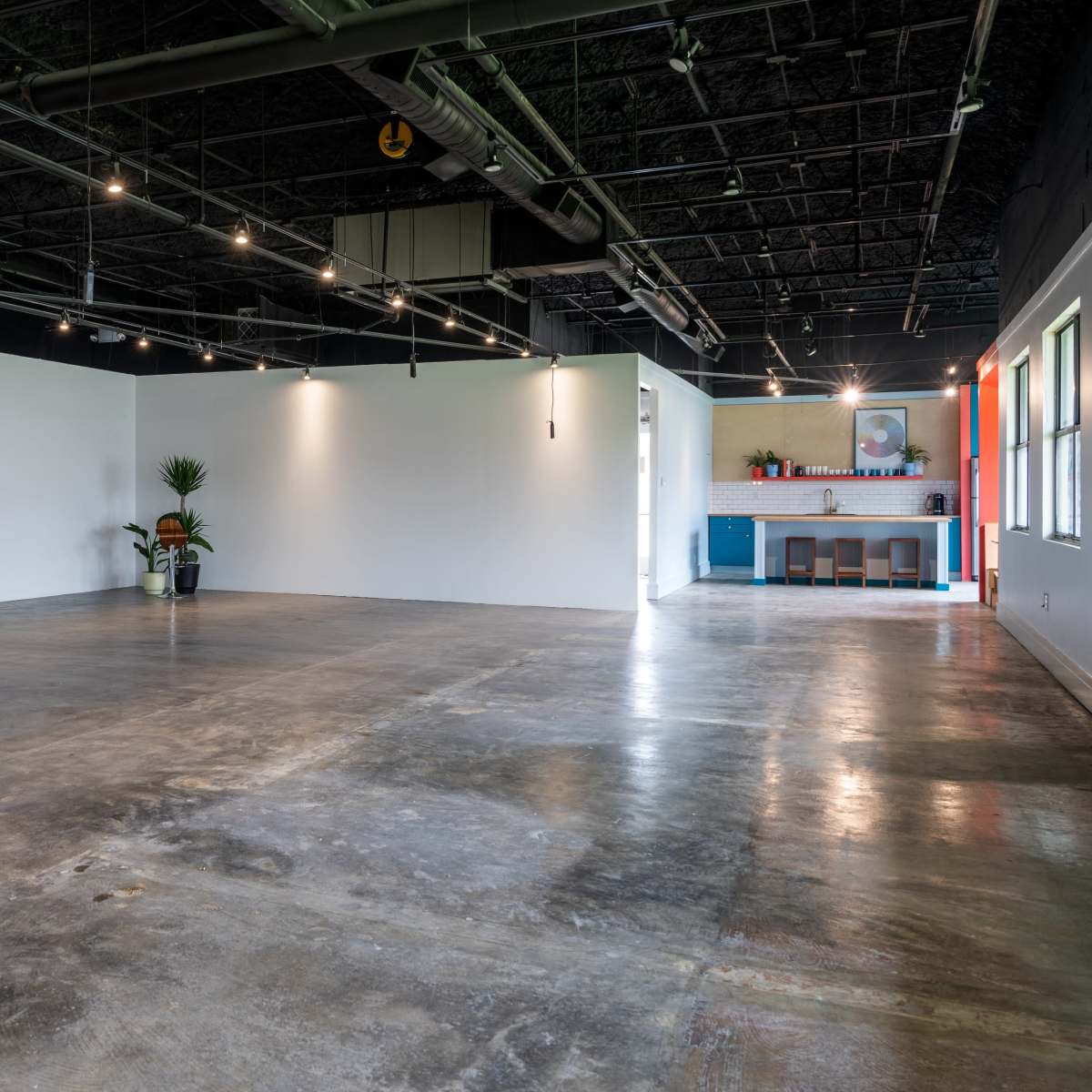 An empty interior space with concrete flooring, a chair and small table against one wall, and a kitchen area visible in the background.