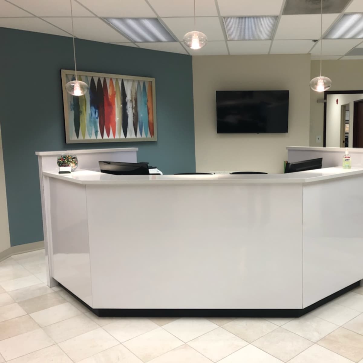 A modern reception area featuring a white front desk, pendant lights, a wall-mounted television, and a colorful artwork in the background.