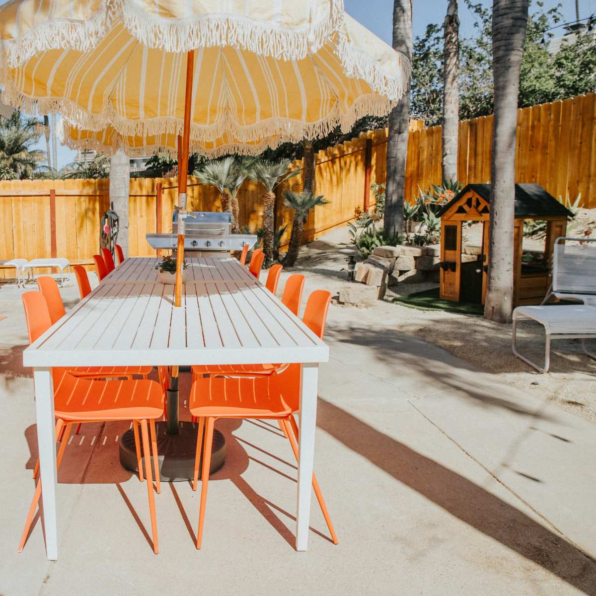 A long white table with orange chairs is set under a large umbrella in a patio area surrounded by palm trees and a wooden fence.
