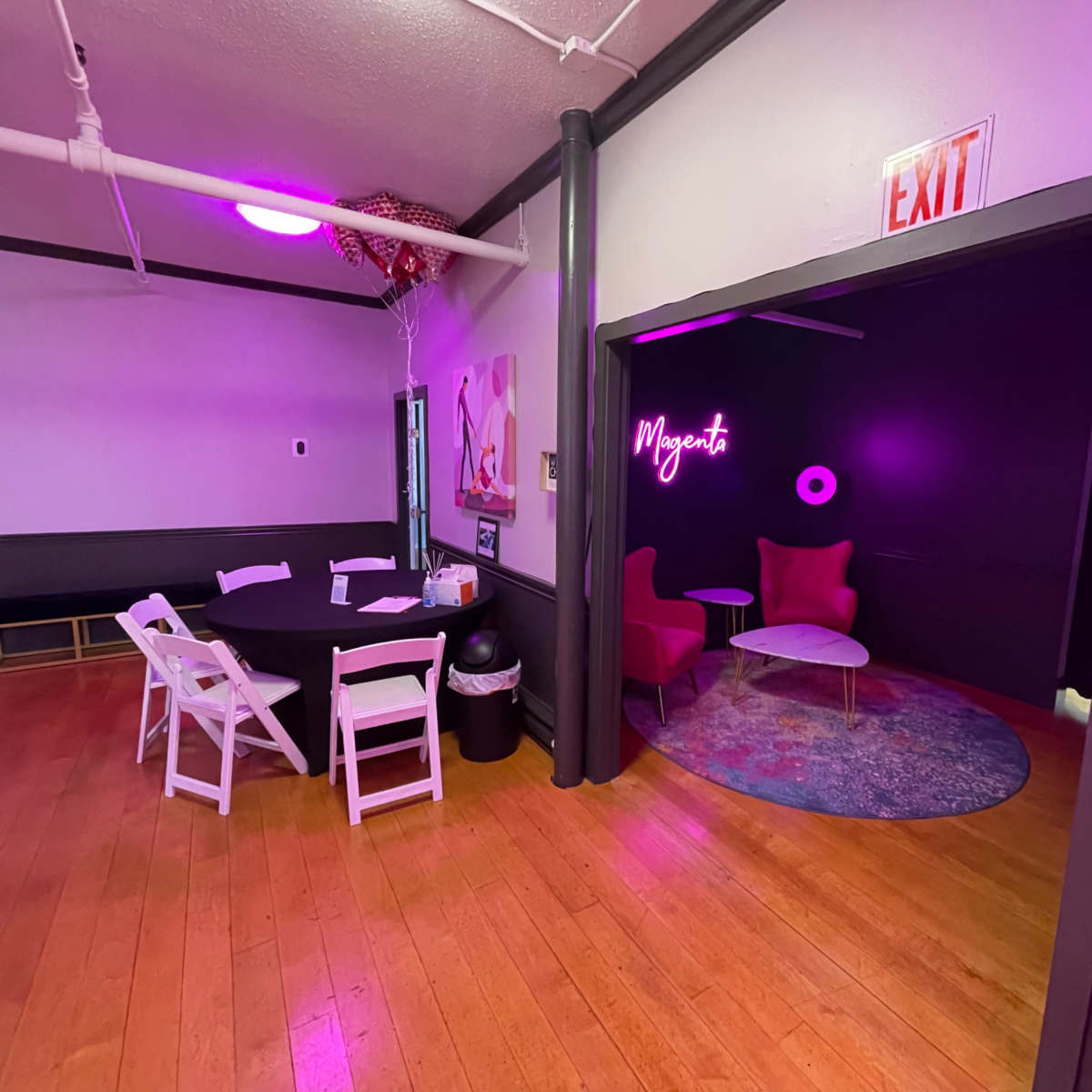 A room with a circular area featuring pink chairs, a small table, and a glowing "Magenta" sign, with a contrasting darker area and a round rug.