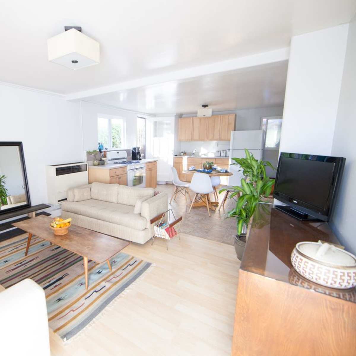 A bright living room with a sofa, a coffee table, a mirrored stand, and a kitchen area in the background.
