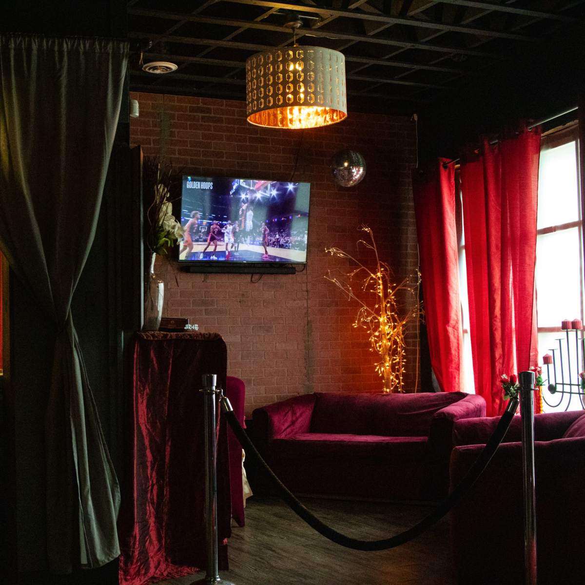 A dimly lit lounge area with a bar, a television displaying a sports event, and plush red seating surrounded by curtains.