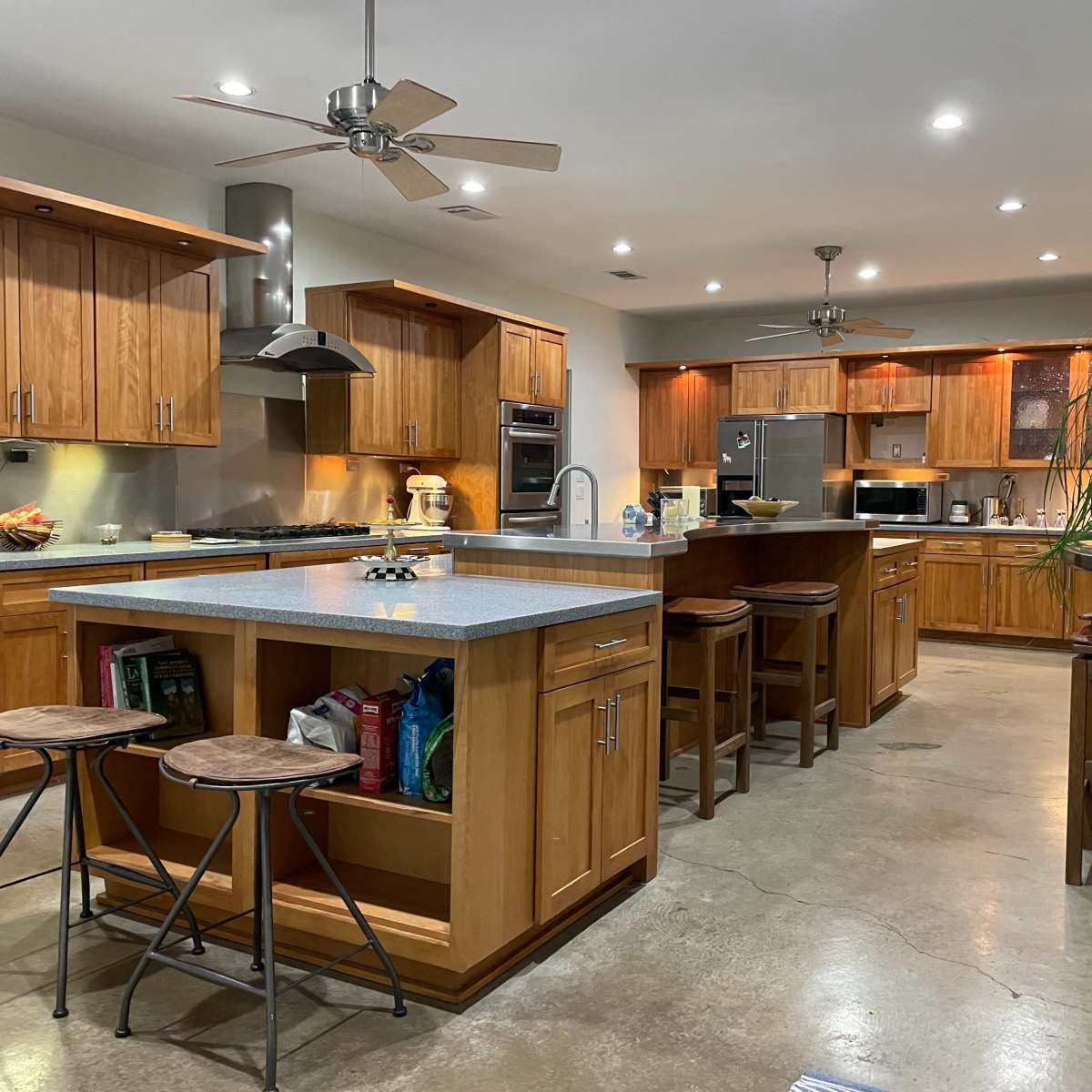 A modern kitchen featuring wooden cabinets, stainless steel appliances, a large island with bar stools, and a concrete floor.