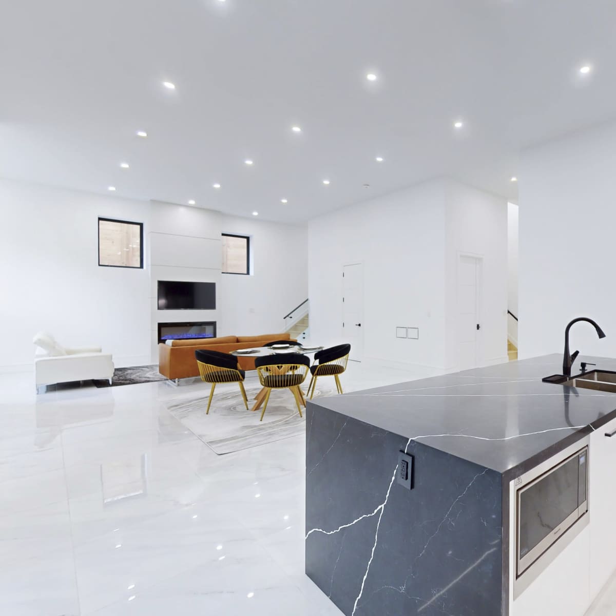 A modern, open-concept kitchen and living area with a large island, sleek appliances, and a bright, airy atmosphere created by white walls and marble flooring.