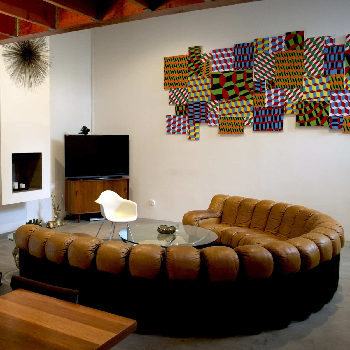 A modern living room featuring a curved brown leather sofa, a glass coffee table, a television, and colorful abstract wall art.